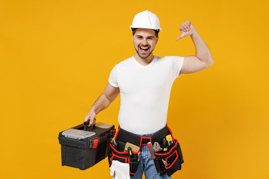 Young smiling employee handyman man in protective helmet hardhat point thumb finger on tool case box isolated on yellow background Instrument accessories renovation apartment room Repair home concept