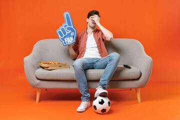 Young man football fan in shirt support team with soccer ball sit on home sofa watch tv live stream hold pizza foam glove finger do facepalm gesture isolated on orange background People sport concept.