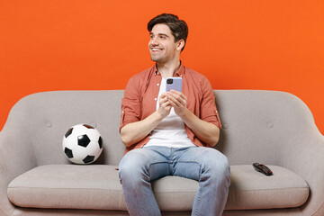 Young man football fan in shirt support team with soccer ball sitting on sofa home watch tv live...