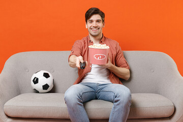 Young fun man football fan in shirt support team with soccer ball sit on sofa home watch tv live stream hold takeaway pop corn bucket switch channel isolated on orange background People sport concept.