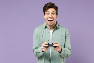 Young fun cheerful excited happy man 20s wearing casual green mint shirt white t-shirt playing pc game with joystick console isolated on purple color background studio portrait. Lifestyle concept