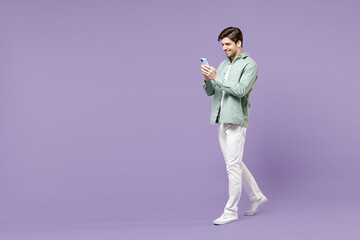 Full length young fun happy man in casual mint shirt white t-shirt using mobile cell phone chat online in social network walk isolated on purple background studio portrait People lifestyle concept.