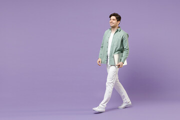 Fototapeta premium Full length young side view smiling man in casual mint shirt white t-shirt hold closed laptop pc computer chat online walk go isolated on purple background studio portrait People lifestyle concept