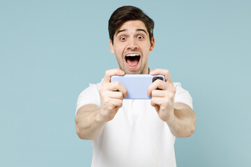 Fototapeta premium Young excited amazed happy man 20s in white casual basic t-shirt using play racing on mobile cell phone hold gadget smartphone for pc video games isolated on pastel blue background studio portrait