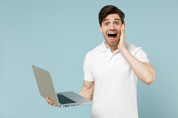 Young shocked surprised man in white casual basic t-shirt hold laptop pc computer chat online browsing internet hold face isolated on pastel blue background studio portrait People lifestyle concept.