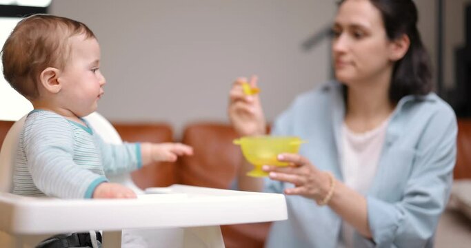Young mother feeding her one year son with a spoon in baby chair at home. Motherhood and baby food concept