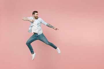 Full length young excited unshaven man wear blue striped shirt white t-shirt jump high point index finger aside on copy space workspace area isolated on pastel pink background. Tattoo translate fun