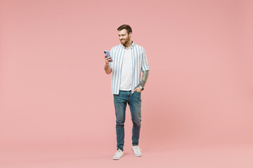 Full length young smiling happy unshaven man 20s in blue striped shirt holding mobile cell phone chat online browsing internet isolated on pastel pink background studio portrait. Tattoo translate fun