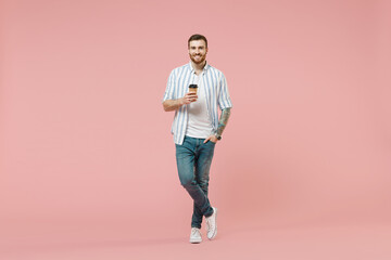 Fototapeta na wymiar Full length of young happy unshaven man 20s wear blue striped shirt hold takeaway delivery craft paper brown cup coffee to go isolated on pastel pink background studio portrait. Tattoo translate fun