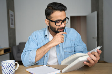 Young indian latin eastern male student in glasses reading book writing notes in textbook at home...