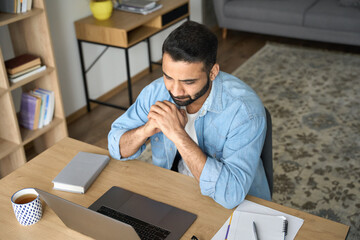 Young adult latin student guy watching online virtual training webinar using pc laptop sitting at modern home office. Top view of indian Hispanic man looking at computer screen on desk indoors.