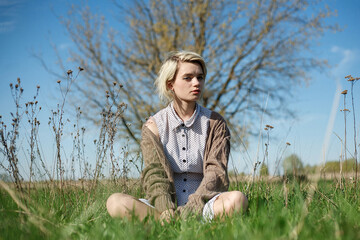 Naklejka na ściany i meble young attractive caucasian female with short hair in spotted dress and sweater sitting on a ground at field on sunny day