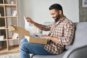 Happy smiling latin indian man opening box with ordered goods at home on couch. Online shopper male...