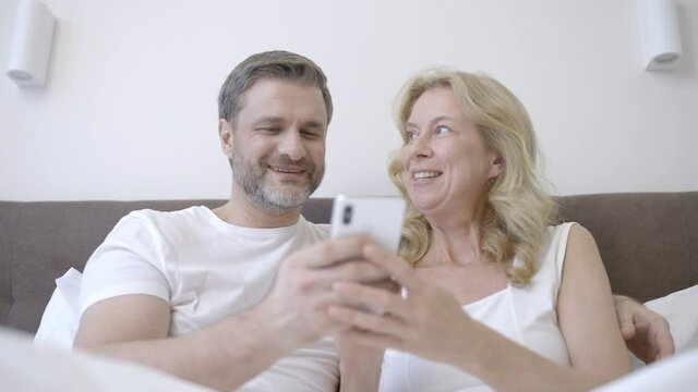Happy middle-aged loving couple scrolling smartphone together, resting in bed