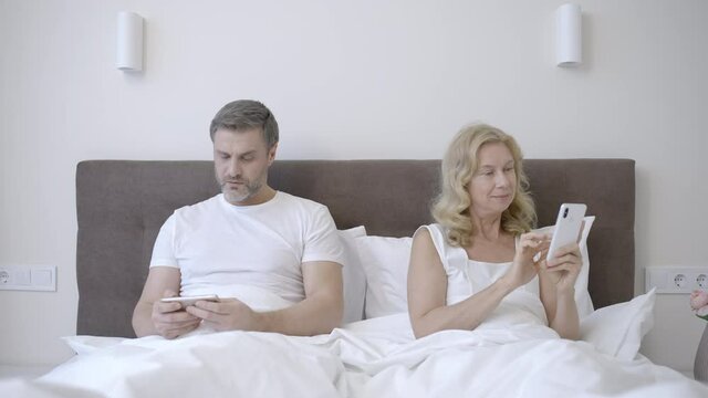 Middle-aged couple using gadgets in bed, scrolling phones, relationship crisis