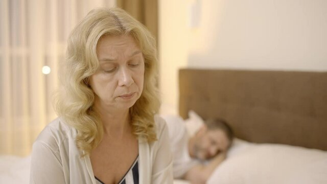 Sad mature woman sitting in bed, looking at sleepy husband, unhappy marriage