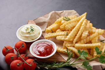 Homemade baked potato fries with mayonnaise, tomato sauce and rosemary on wooden board. tasty french fries on cutting board, in brown paper on black stone table background, unhealthy food.
