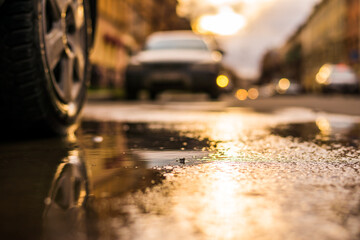 Streets in the light of the autumn sun after the rain, view from under the wheels of a parked car. View from puddles on the pavement level