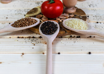 Spices, peppercorns, coriander berries and rice in three wooden spoons, prepared for a traditional meal, placed on a wooden background in the middle of which is a tomato