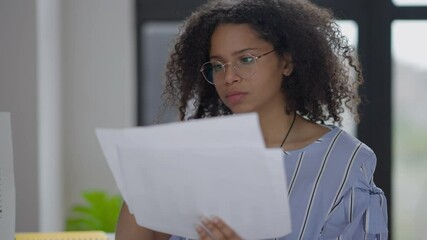 Portrait of thoughtful young woman in eyeglasses analyzing business graphs sitting in home office indoors. Serious African American analyst working in the morning examining documents