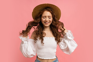 Dreamy  Woman  with close eyes in summer outfit plays hair on pink background. Straw hat, white top with sleeves, natural make up.