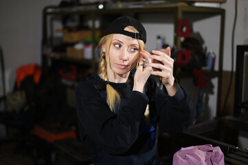 Portrait of a beautiful caucasian blonde girl applies cosmetics, posing in the garage in work clothes
