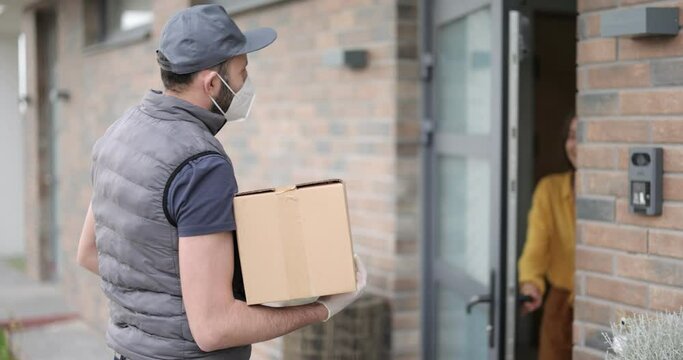 Courier in face mask delivers parcel for a young woman at the doorway of the house. Deliverying goods during a pandemic and happy customer concept