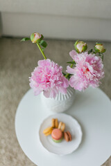 Living room interior. Bouquet of pink peonies in a vase, multicolored French macaroon pastries. Cozy. Spring.
