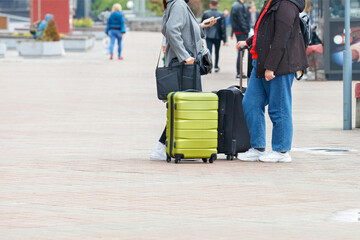 A couple of travelers with suitcases on wheels stand on the sidewalk checking their way on their smartphone.