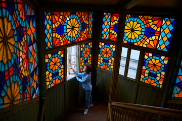 Fototapeta na wymiar Cute brunette girl posing on authentic balcony of an old residential building with a stained glass window made of multicolored mosaics