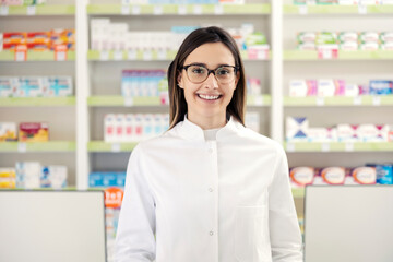Portrait of a pharmacist in a pharmacy. A close-up shot of a pharmacist woman in uniform and with glasses standing in the middle of the pharmacy in front of a medicine shelf. Happy to do the job