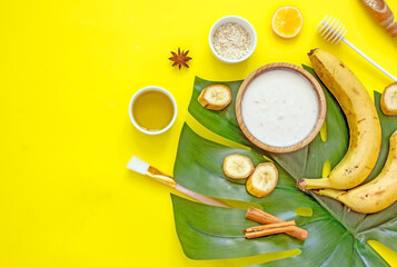 Ingredients for a natural cosmetic facial mask with your own hands. Banana pulp, honey, butter, yolk, oatmeal on a yellow background with exotic leaves, top view, flat lay, copy space. 