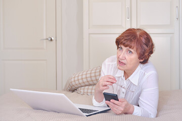 Senior woman wearing glasses, using mobile phone, laptop on the bed at home.