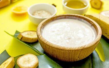 Ingredients for a natural cosmetic facial mask with your own hands. Banana pulp, honey, butter, yolk, oatmeal on a yellow background with exotic leaves, top view, flat lay, copy space. 