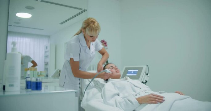 A female client is laying in the cosmetology chair in an apron while a female cosmetologist is doing a procedure non-injection mesotherapy apparatus on her cheek by using the Dermadrop DTA device.