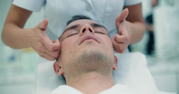 Close-up face of a male client is laying in the chair while a female cosmetologist is doing a massage of his cheeks and temples.