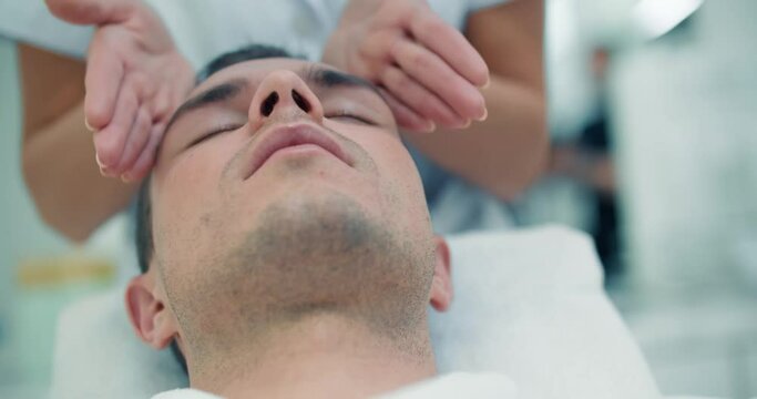 Close-up face of a male client is laying in a cosmetology bed while a female cosmetologist is slowly doing a massage of his forehead and cheeks.