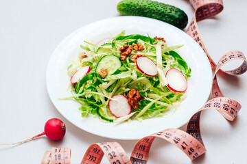 Cucumber salad with radishes, cabbage and nuts on a white plate and a centimeter. Top view on gray background.