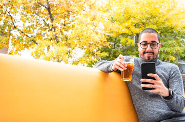 young Latino man with glasses sitting on a piece of furniture outside a bar, drinking beer and checking his phone. - Powered by Adobe