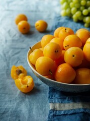 Fresh fragrant yellow plums in a cup on a blue linen tablecloth, summer still life. Green grapes in the background. Bright sunshine on berries. Copy space. Complementary colors: yellow and blue.