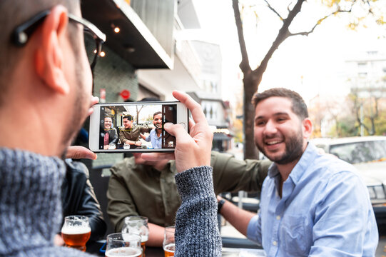 boy taking a picture of a group of friends sitting at a table drinking beer outside a bar.