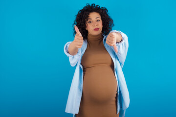 young Arab pregnant woman wearing dress against blue wall showing thumbs up and thumbs down,...