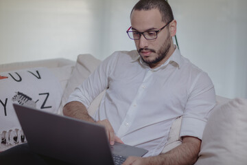 Joven latino trabaja online con su laptop desde la comodidad de su sofá, navegando en internet, y viendo noticias en portales digitales
