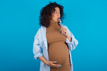 Very hungry young Arab pregnant woman wearing dress against blue wall holding spoon into mouth...