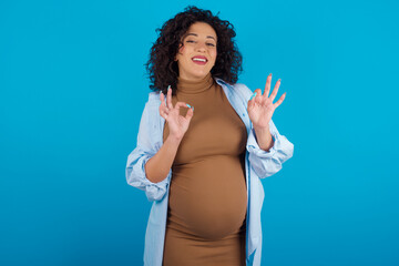 young Arab pregnant woman wearing dress against blue wall showing both hands with fingers in OK sign. Approval or recommending concept