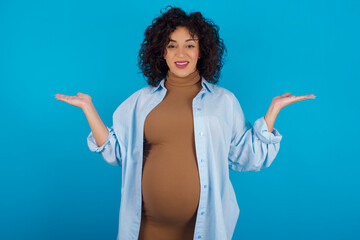 Cheerful cheery optimistic young Arab pregnant woman wearing dress against blue wall holding two...
