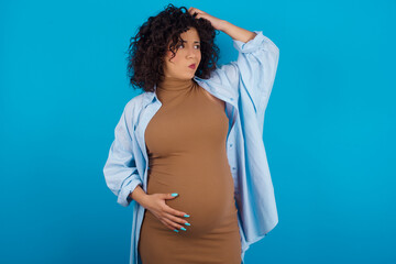 young Arab pregnant woman wearing dress against blue wall confuse and wonder about question....