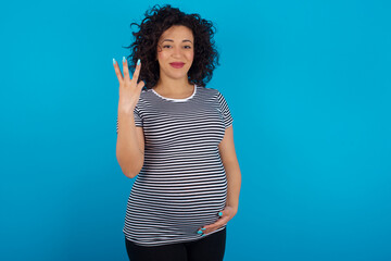 young Arab pregnant woman wearing stripped T-shirt  against blue wall smiling and looking friendly, showing number three or third with hand forward, counting down