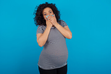 Stunned young Arab pregnant woman wearing stripped T-shirt  against blue wall covers both hands on mouth, afraids of something astonishing