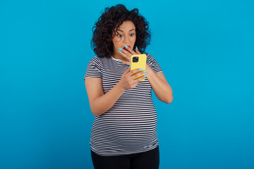 young Arab pregnant woman wearing stripped T-shirt  against blue wall being deeply surprised,...